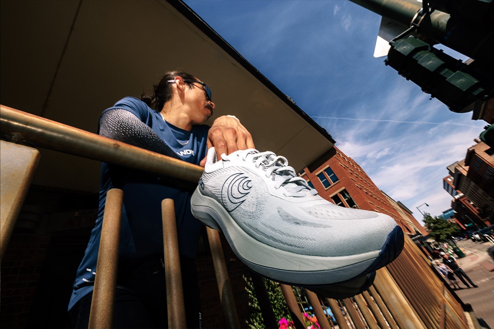 A person holding grey running shoes with a wide toe box while standing on a deck, ready for a comfortable run.” width=